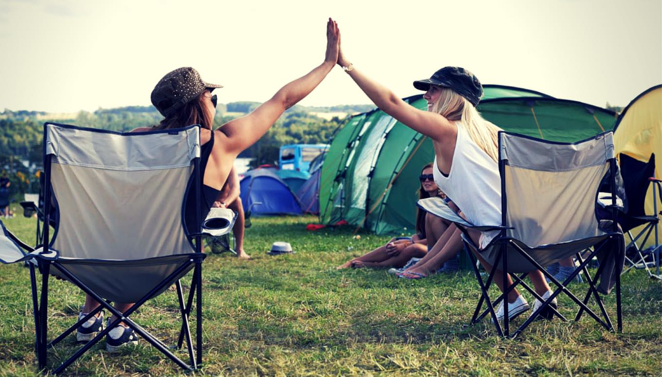 Two friends sat in camp chairs at a festival in their hats and sunglasses