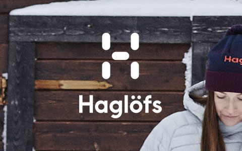 A woman is outside in front of a wooden house, wearing Haglofs gear.