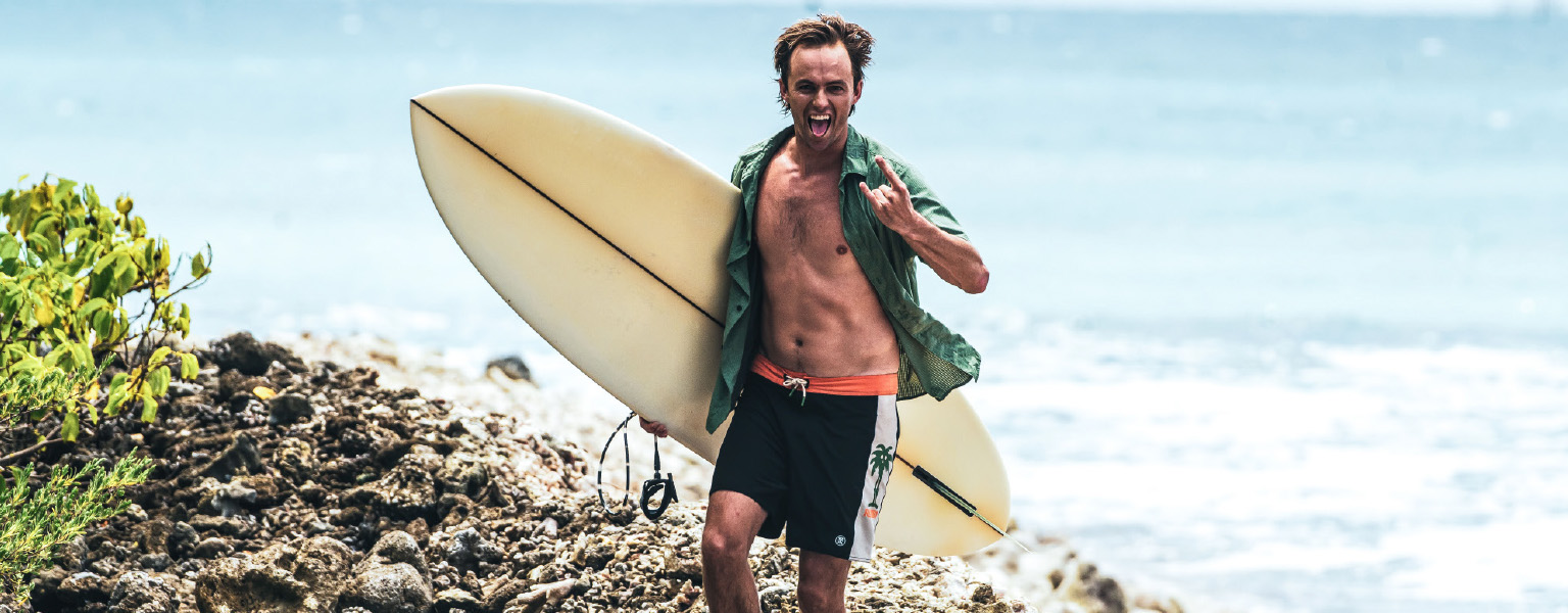 A man is walking away from the sea, carrying a surfboard, wearing Roark shorts and shirt. 