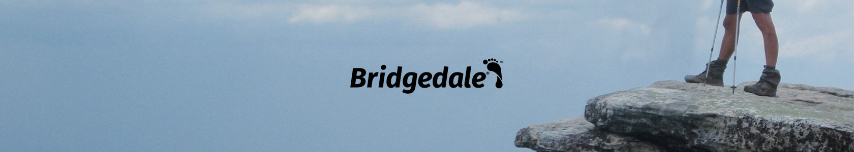 A man is standing on the edge of a big stone, holding trekking poles and wearing Bridgedale socks