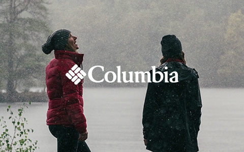 Two people are standing by a lake during falling snow, wearing Columbia clothing.
