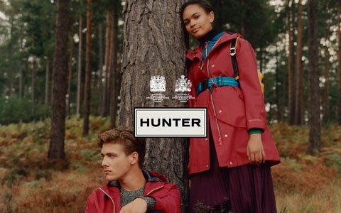 A man sitting by a tree on the ground and a woman standing next to him and the tree, wearing Hunter gear.