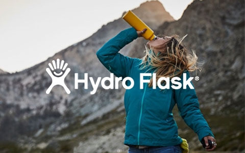 A woman is drinking out of a yellow Hydro Flask bottle somewhere outdoors, surrounded by mountains.