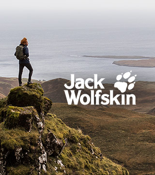 Two people in Jack Wolfskin gear standing by the lake among mountains that are covered by trees and snow.