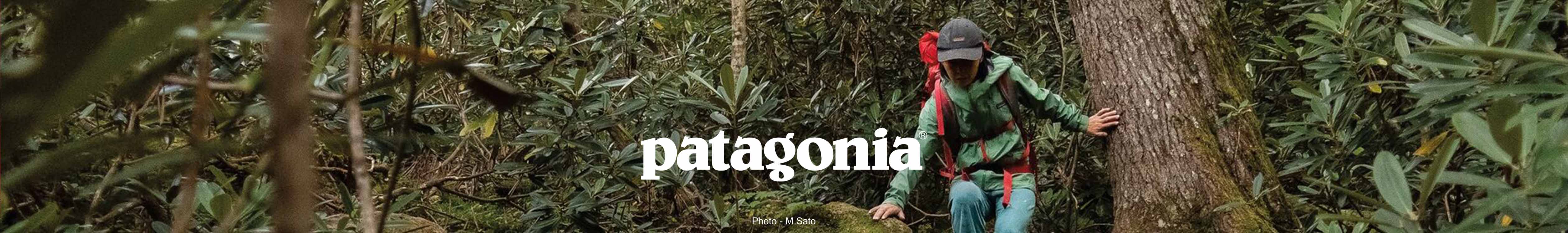 A woman is walking next to a big tree, wearing Patagonia clothes and Patagonia cap, with Patagonia rucksack on her back. 