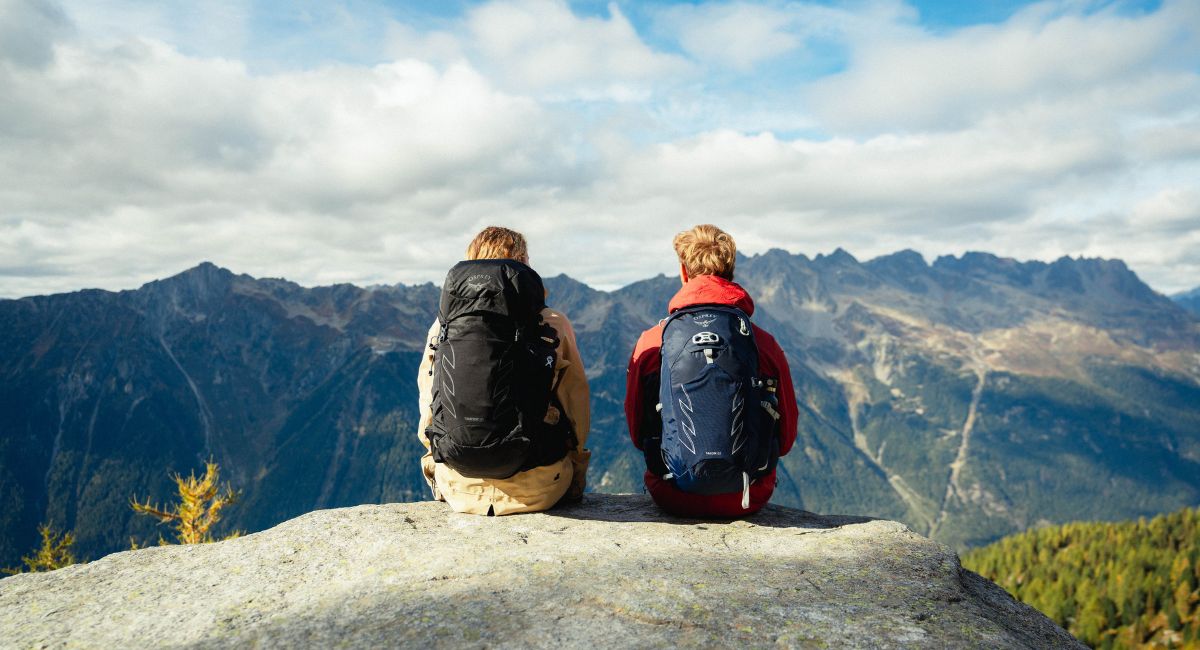 A woman and man with the 70L Fairview and Farpoint backpacks sit looking at a mountain vista in the sunshine with us behind them