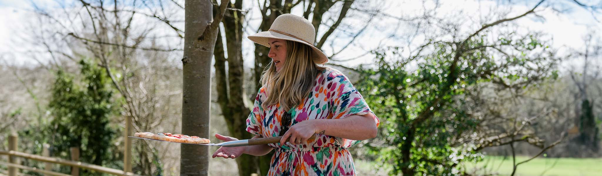 A woman is in a summer dress and in a summer hat, carrying a pizza, while outdoors