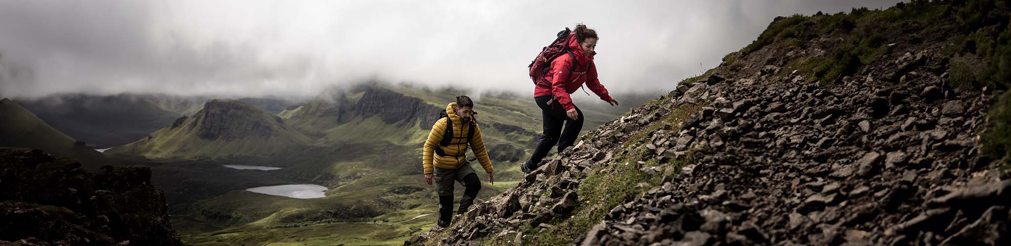 A woman at the front and a man behind her are walking up a steep hill, wearing jackets and rucksacks.