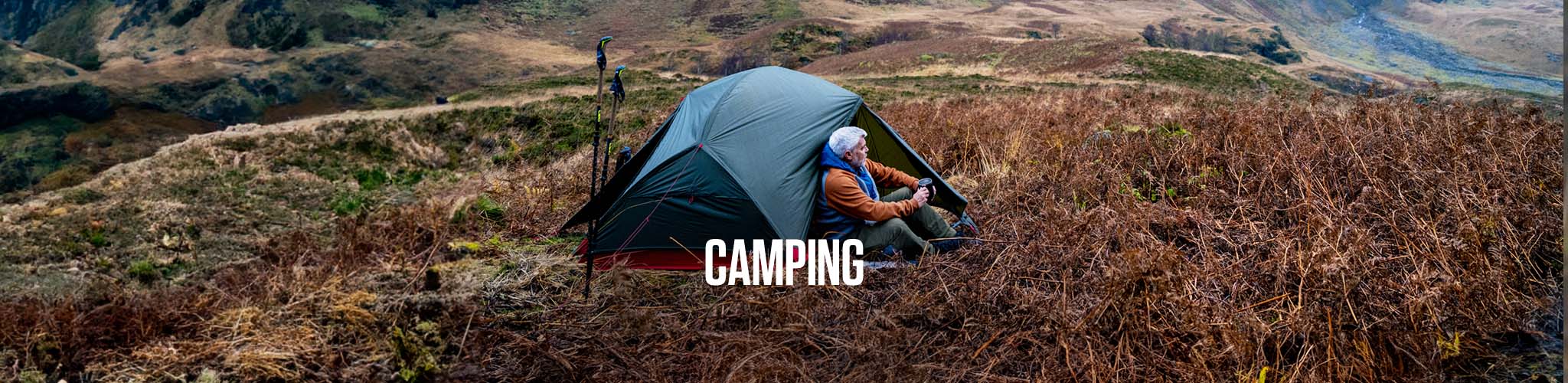 A man is sittng with his back to a tent, dringing from a cup, wearing walking kit