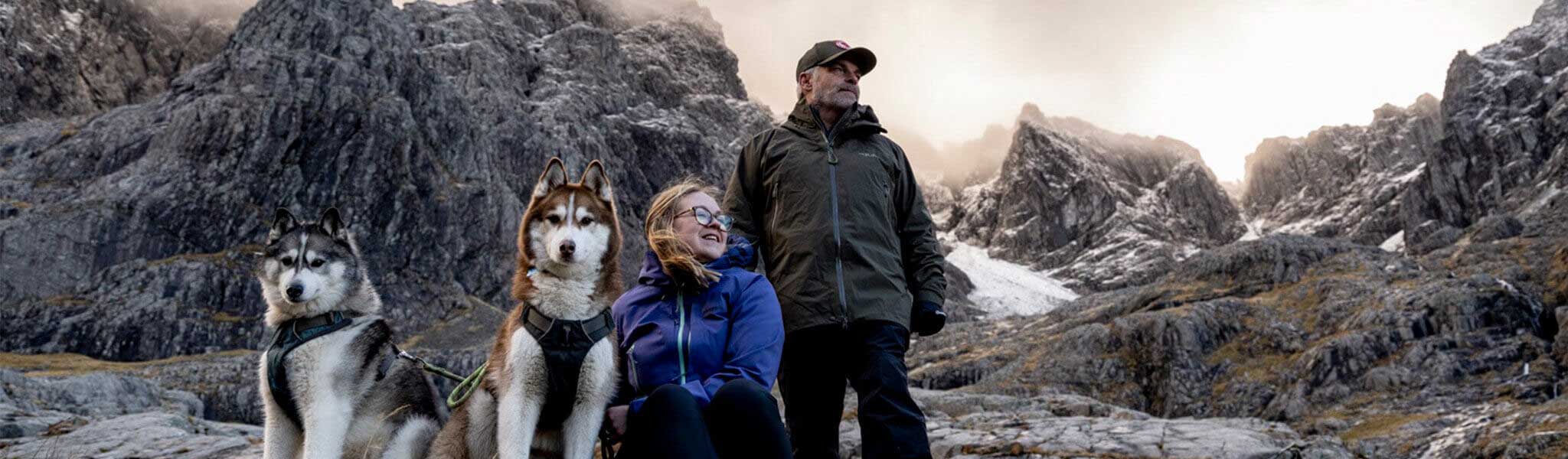 A man standing with a woman sitting next to a sitting dog amongs mountains, wearing walking kit
