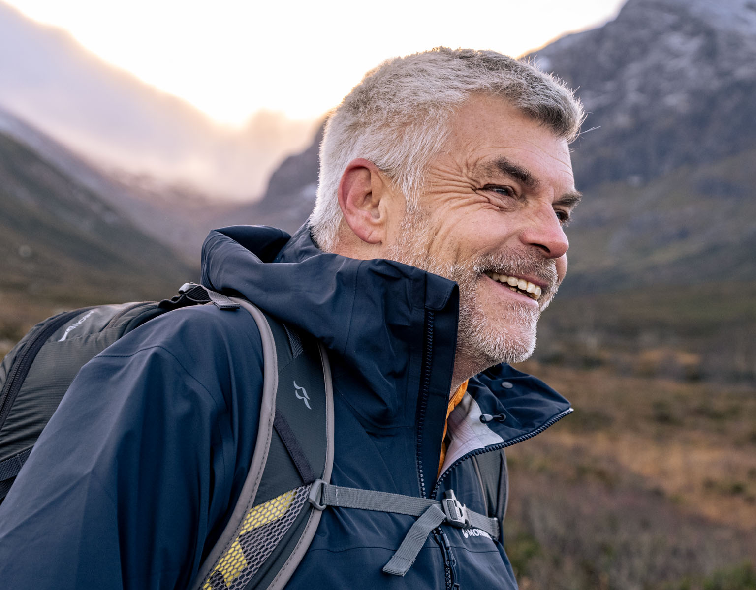 A man is smiling somewhere surraunded by mountains, wearing a blue waterproof jacket and a backback.