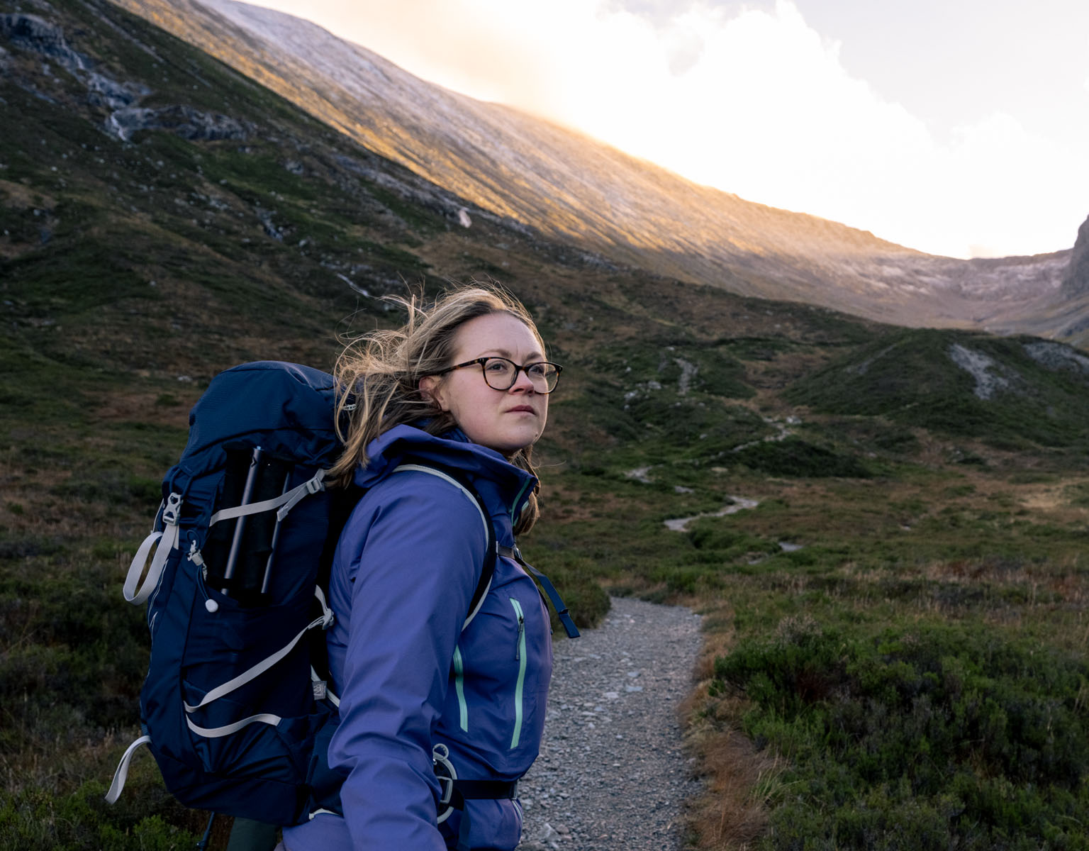 A woman is walking on a foot path somewhere outdoors, wearing a blue waterproof jacket and a rucksack.