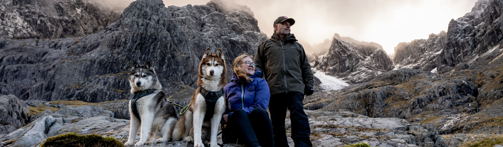 A man and a woman is with two Husky dogs somewhere in the mountains