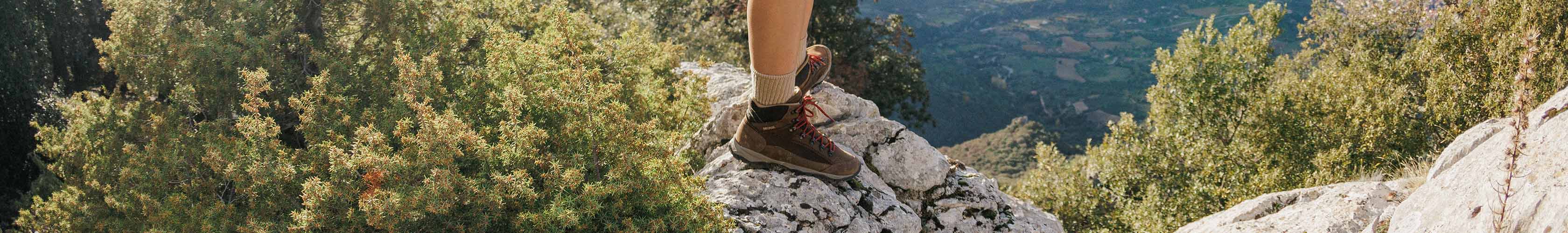 Two pairs of hiking boots worn by two people balance on the edge of some muddy grass.